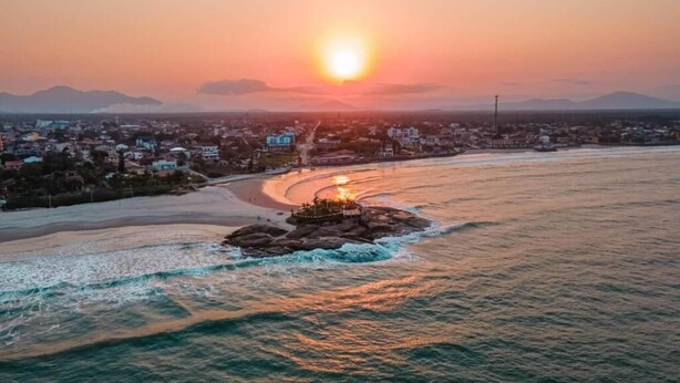 A coastal cityscape at sunset with a beach, ocean, and distant mountains. - Hotel Vila Águamar (Itapoá)