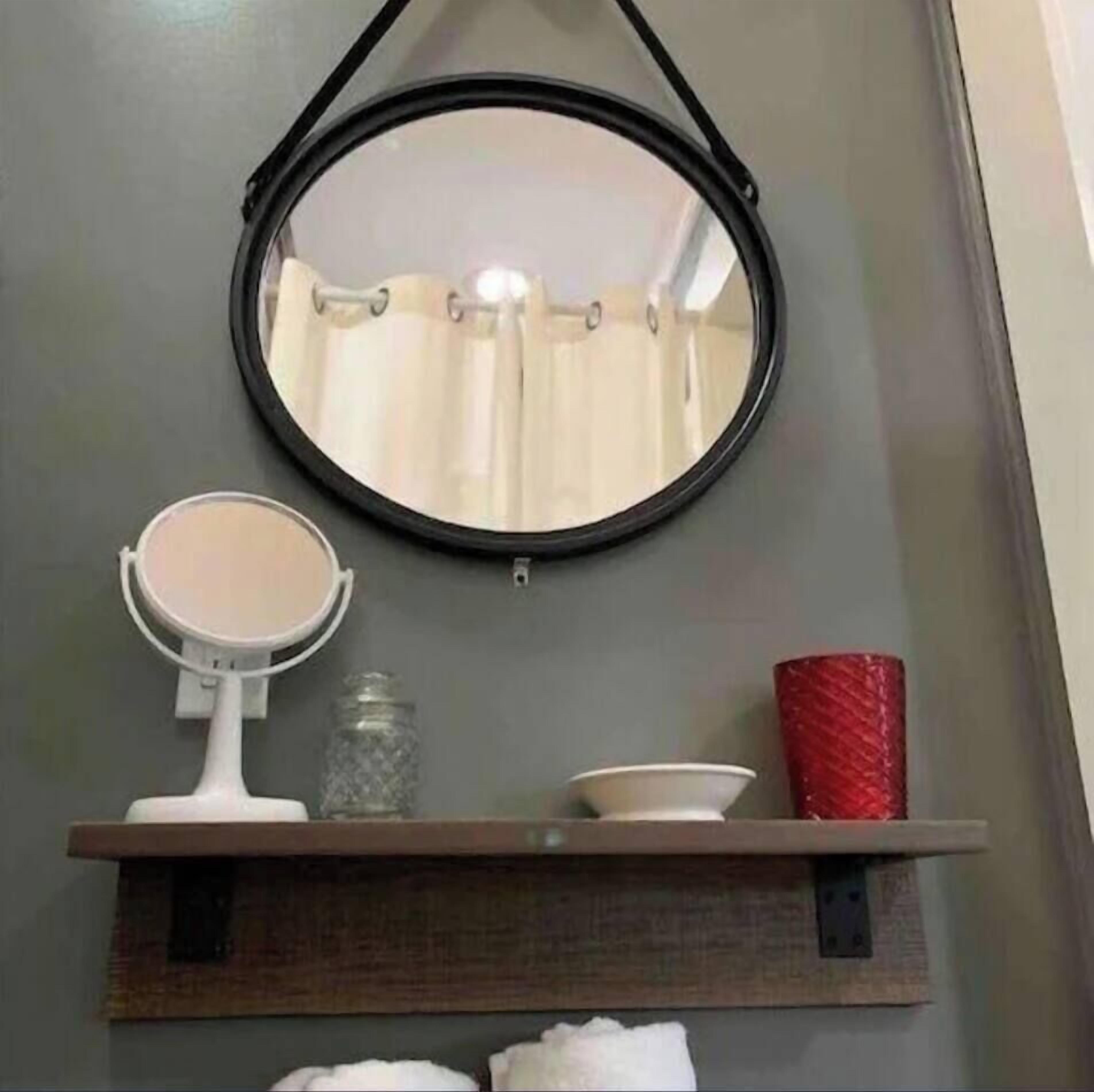 Un baño con un espejo redondo, un espejo más pequeño, un estante con artículos de aseo y una taza roja.