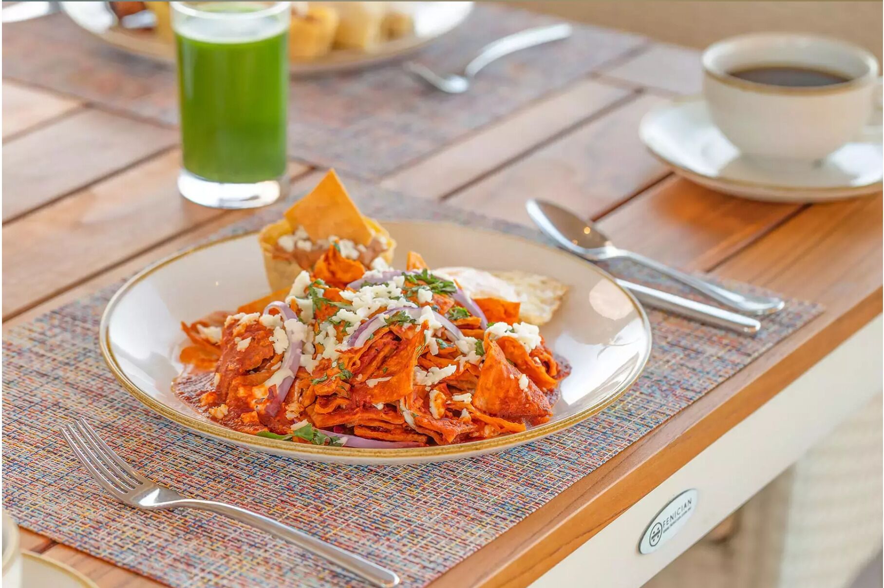 Une assiette de nachos recouverts de fromage et d’oignons rouges est posée sur une table, sur laquelle on trouve également une nappe, une tasse de café et, au fond, une boisson verte.