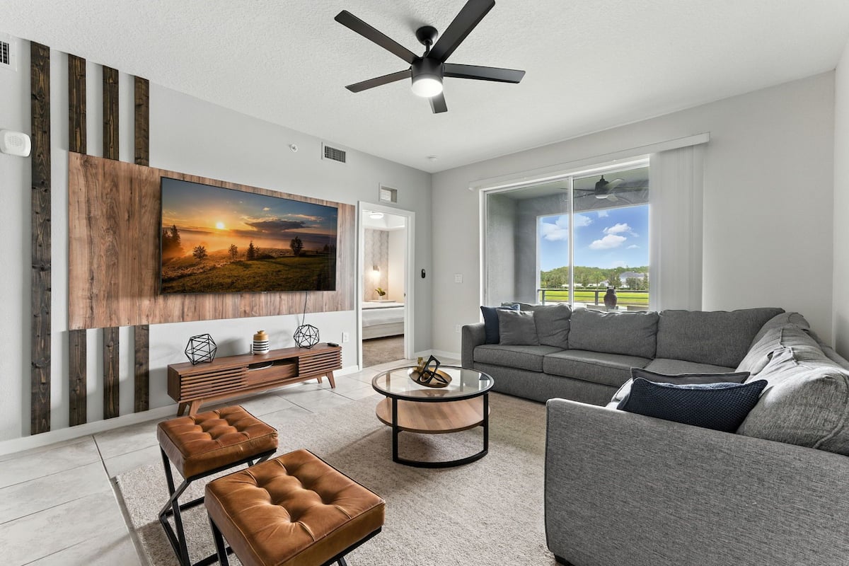 A modern living room with a large flat-screen TV, a grey sofa, a coffee table, and a ceiling fan.