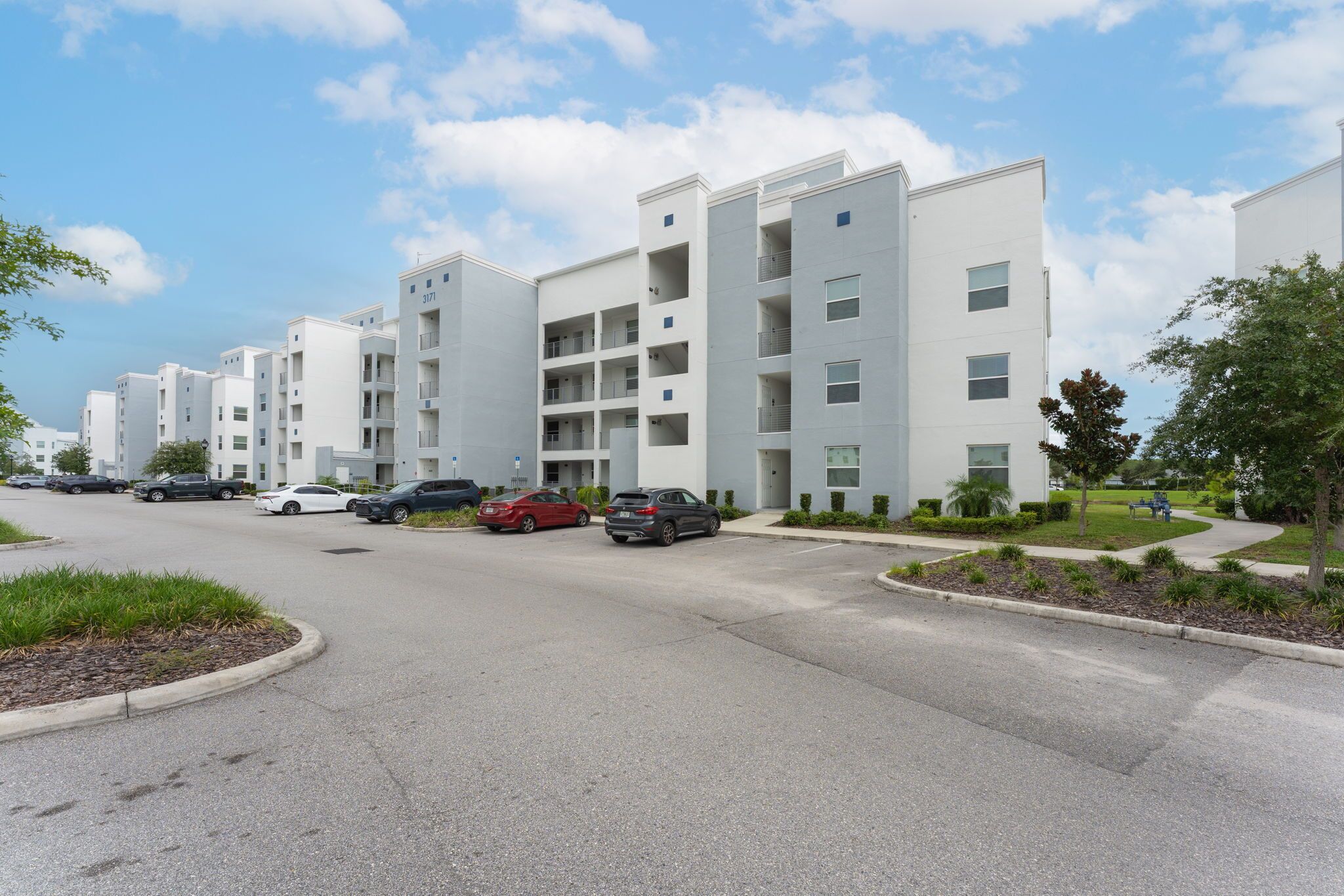 A modern apartment building with multiple floors and balconies, a parking lot with several cars, and a well-maintained landscaped area with trees and shrubs.