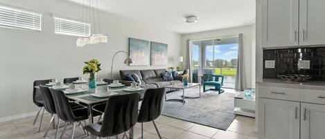 A modern kitchen with white cabinets, a dining area with a table and chairs, and a living room with a sofa and a view of the outdoors.