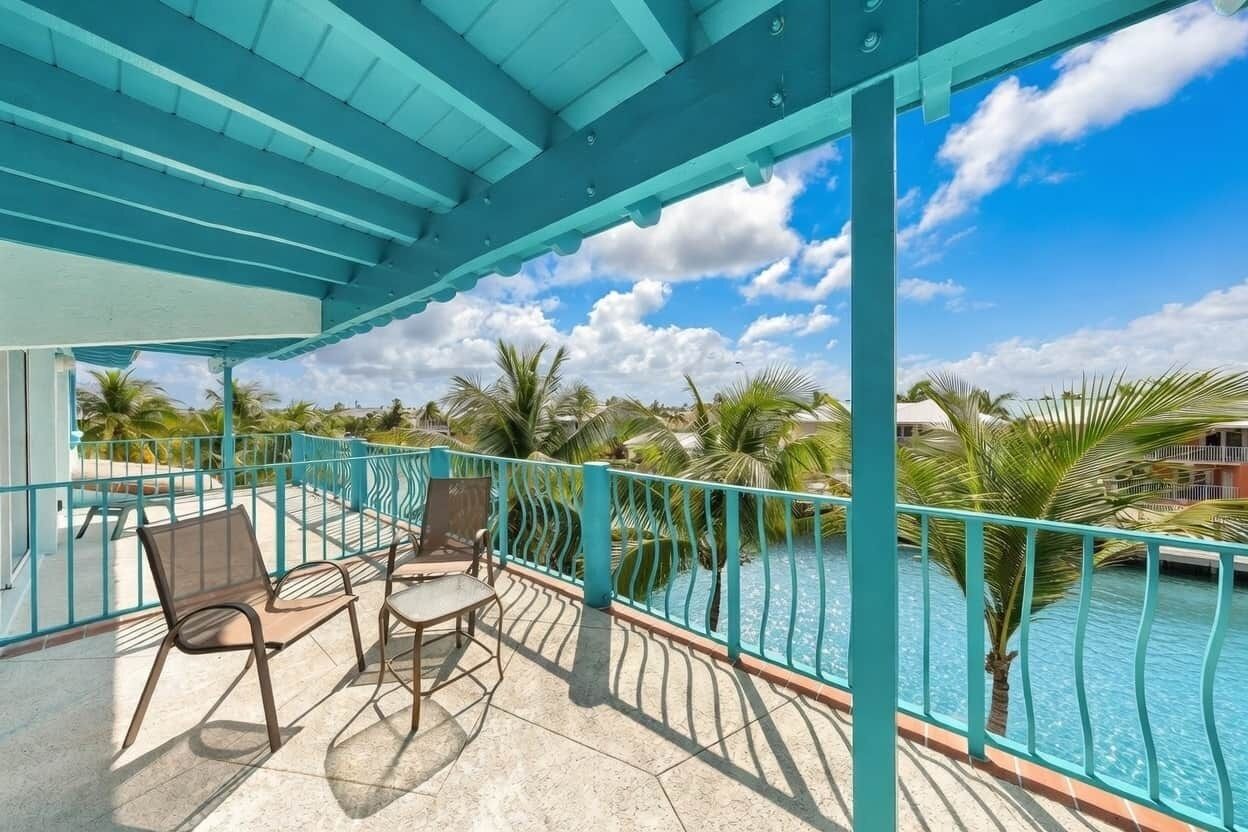 Un balcón techado con vista a la piscina, barandilla turquesa y mobiliario exterior.