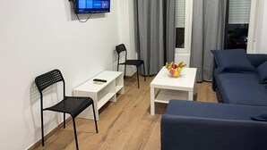 Una sala de estar moderna con un televisor de pantalla plana montado en la pared, una mesa de centro blanca, dos sillas negras, un sofá azul y una ventana con cortinas grises.