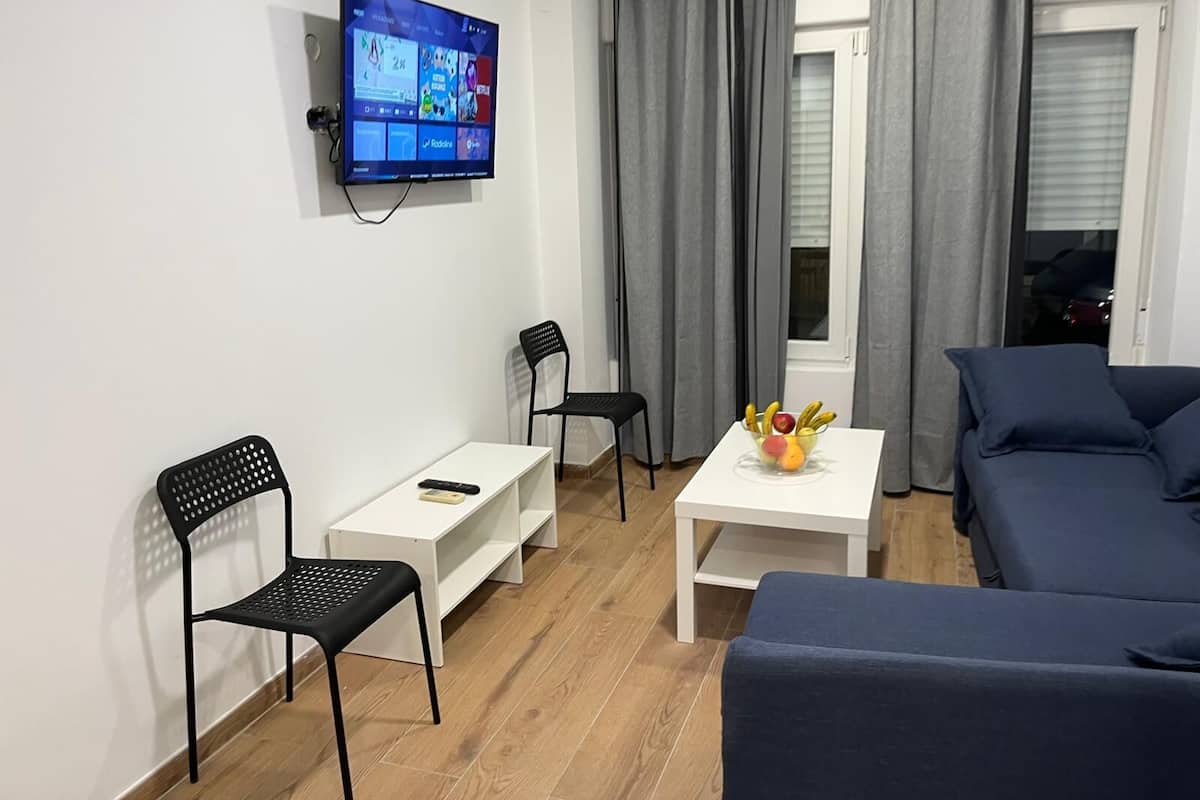 Una sala de estar moderna con un televisor de pantalla plana montado en la pared, una mesa de centro blanca, dos sillas negras, un sofá azul y una ventana con cortinas grises.