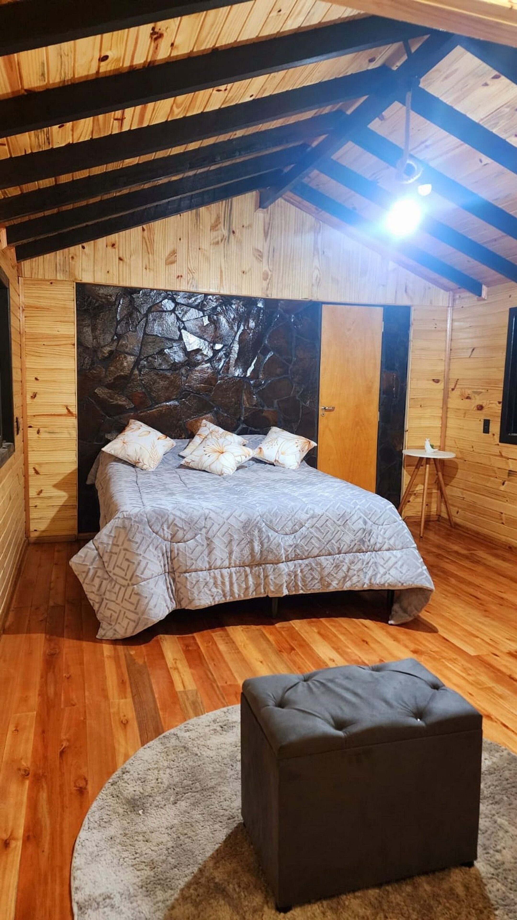 A bedroom with a stone wall, wooden beams, a bed with patterned bedding, and a small ottoman.