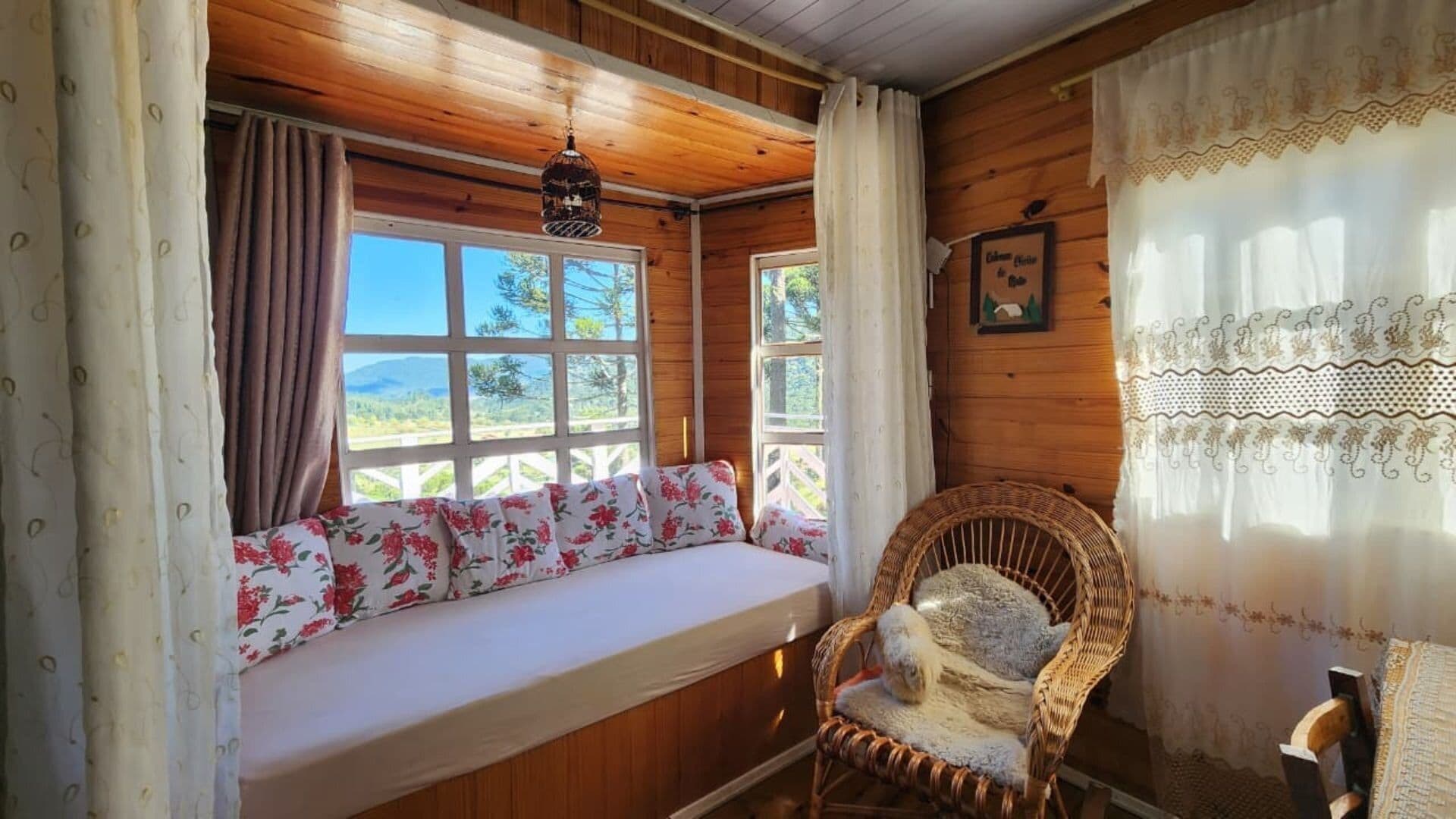 A cozy cabin room with a wooden interior, a bench with floral cushions, a wicker chair, and a view of the outdoors through large windows.