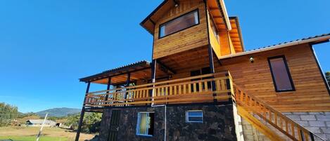 A modern two-story wooden house with a stone foundation, a balcony, and a view of the surrounding landscape.