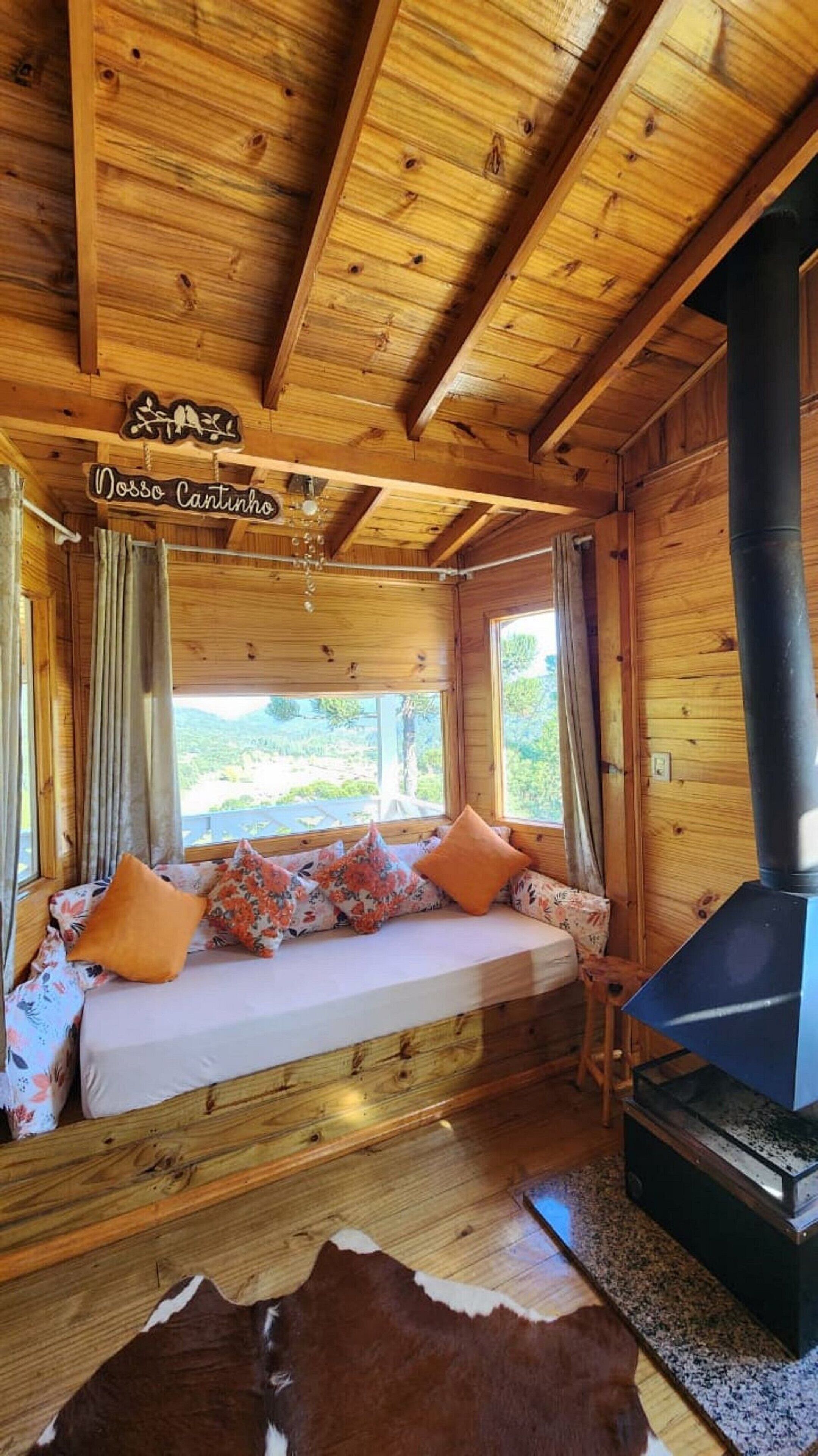 L’intérieur chaleureux d’un chalet avec un plafond en bois, un canapé agrémenté de coussins à motifs floraux, une grande fenêtre et un poêle à bois.