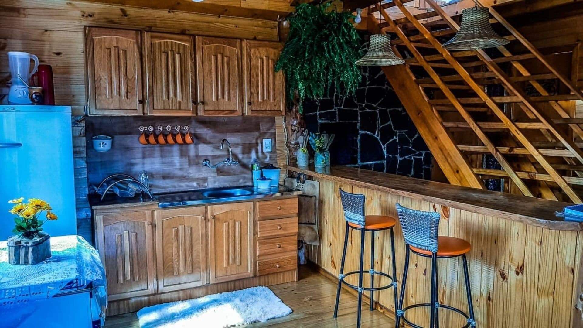 A rustic kitchen with wooden cabinets, a stone wall, and a wooden staircase.
