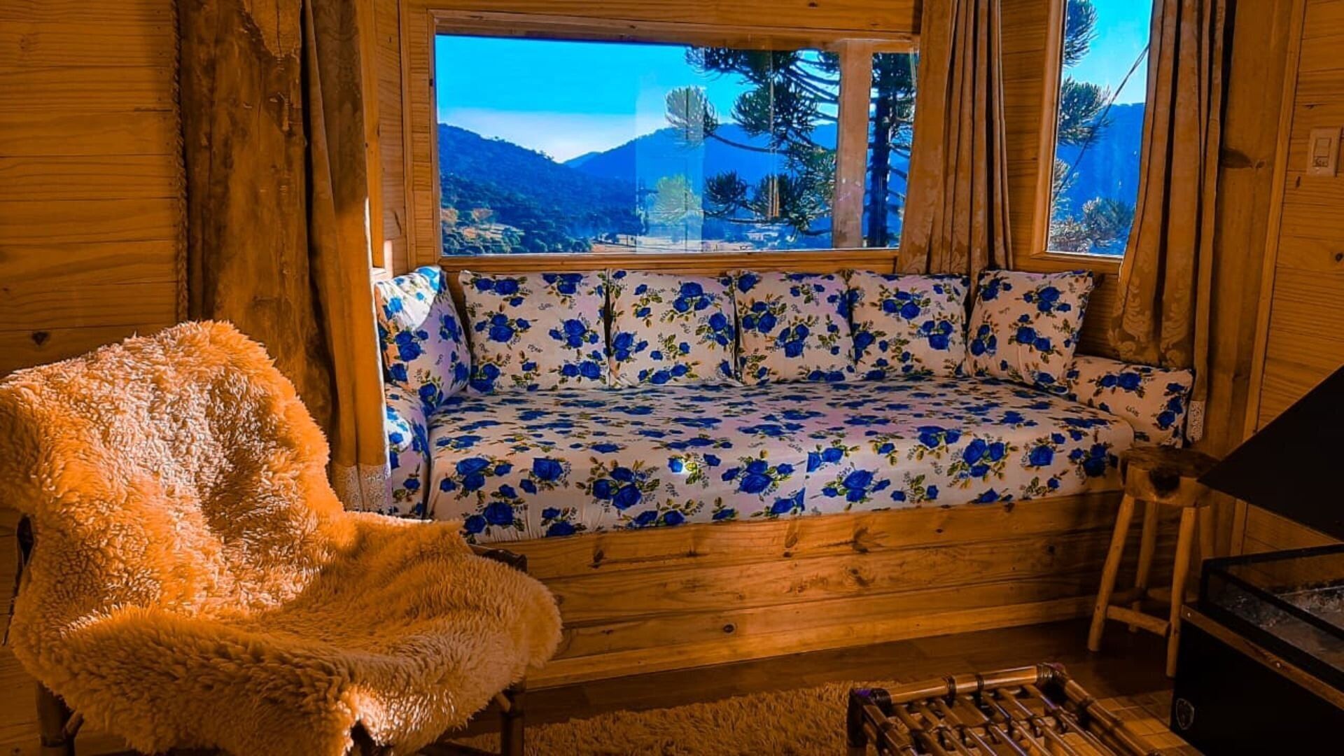 A cozy cabin interior with a wooden bench, a sofa with floral upholstery, and a view of mountains through a window.