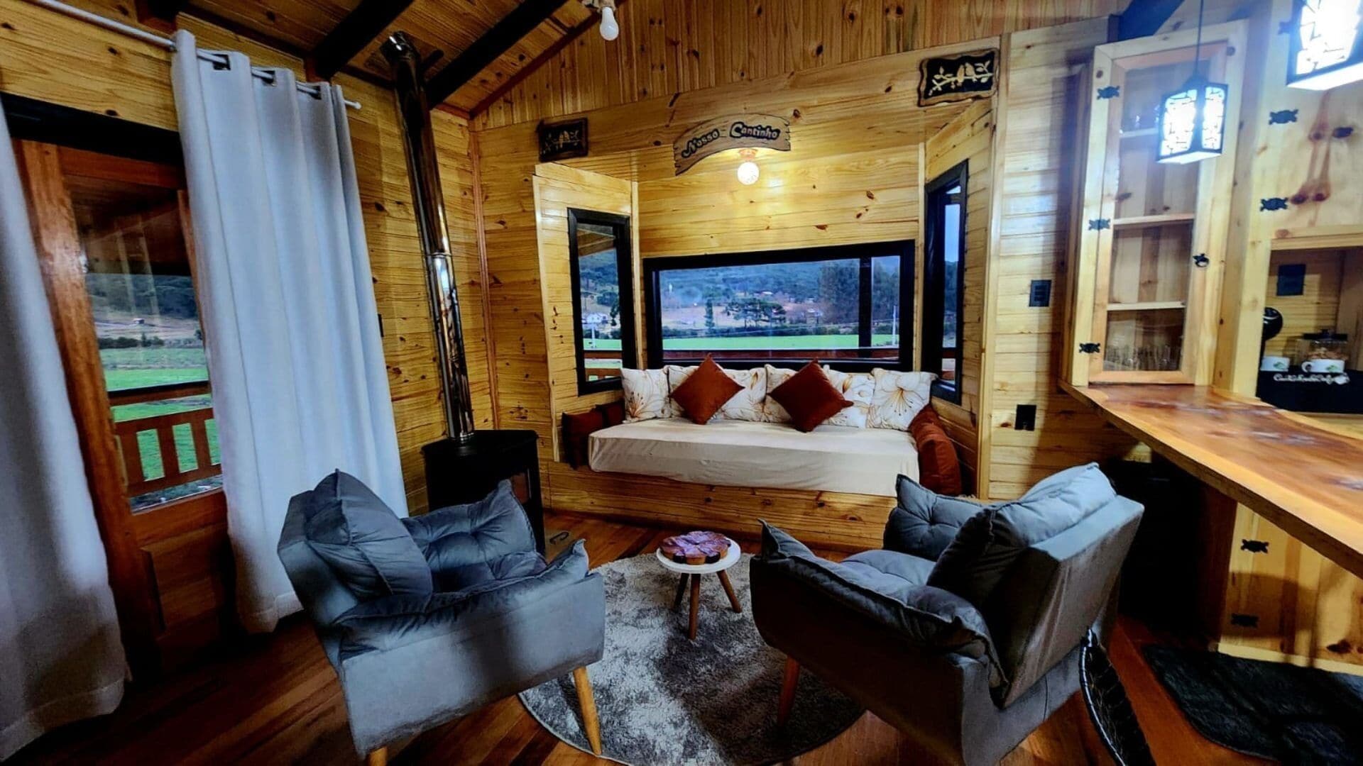Interior acogedor de una cabaña con una cama, dos sofás, una mesita y un ventanal con vistas panorámicas.
