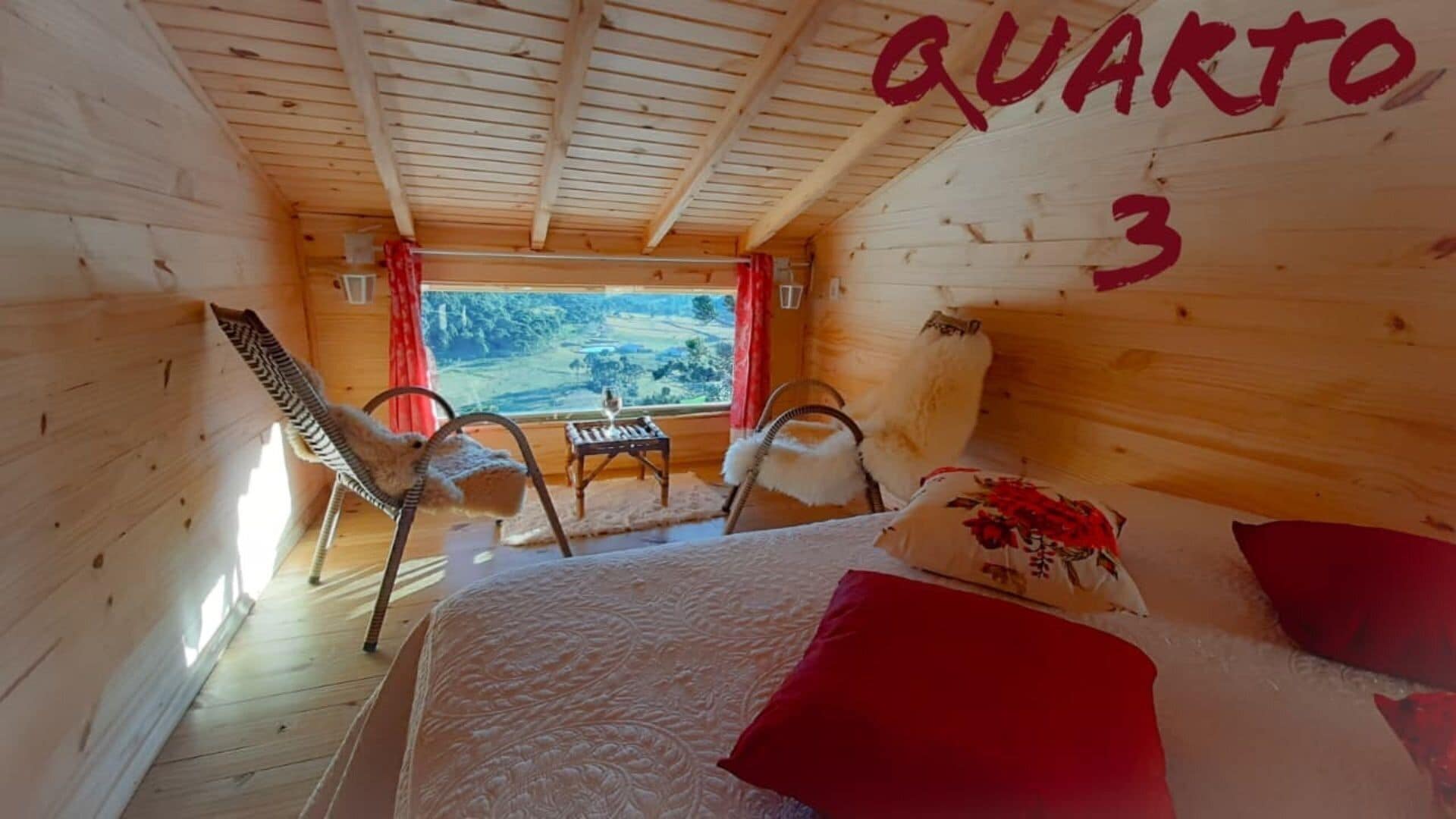 Ein Zimmer mit Holzboden, einem Bett, zwei Stühlen, einem kleinen Tisch mit einem Glas und einem Fenster mit Blick ins Freie. Auf der Wand ist der Text „QUARTO 3“ zu sehen.