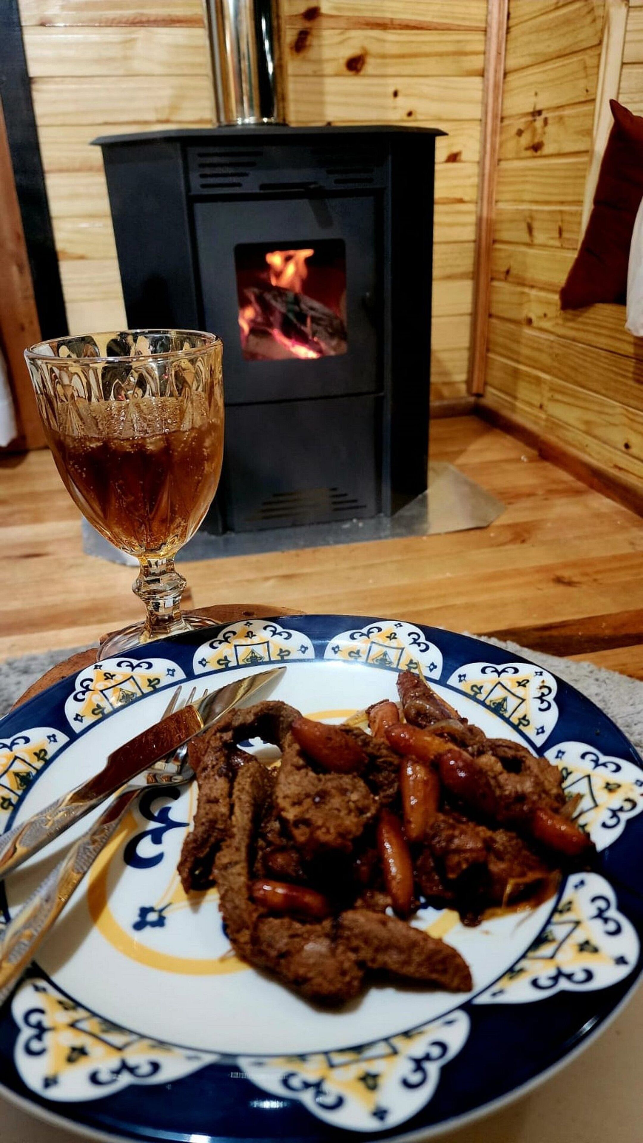 A dining table with a plate of food, a glass of drink, and a wood-burning stove in the background.