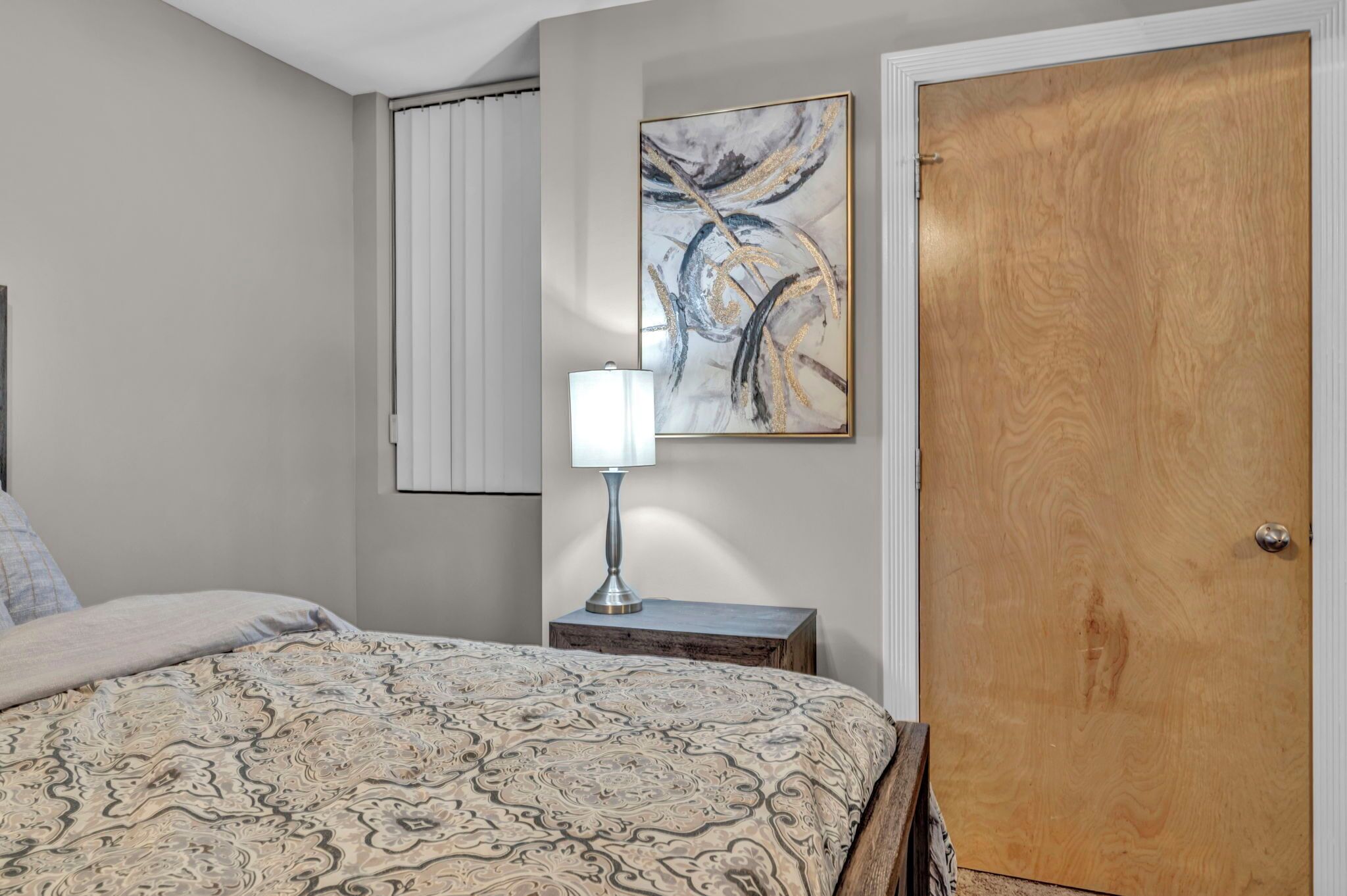 Une chambre à coucher avec un lit, une table de chevet avec une lampe, une porte en bois et un tableau abstrait accroché au mur.