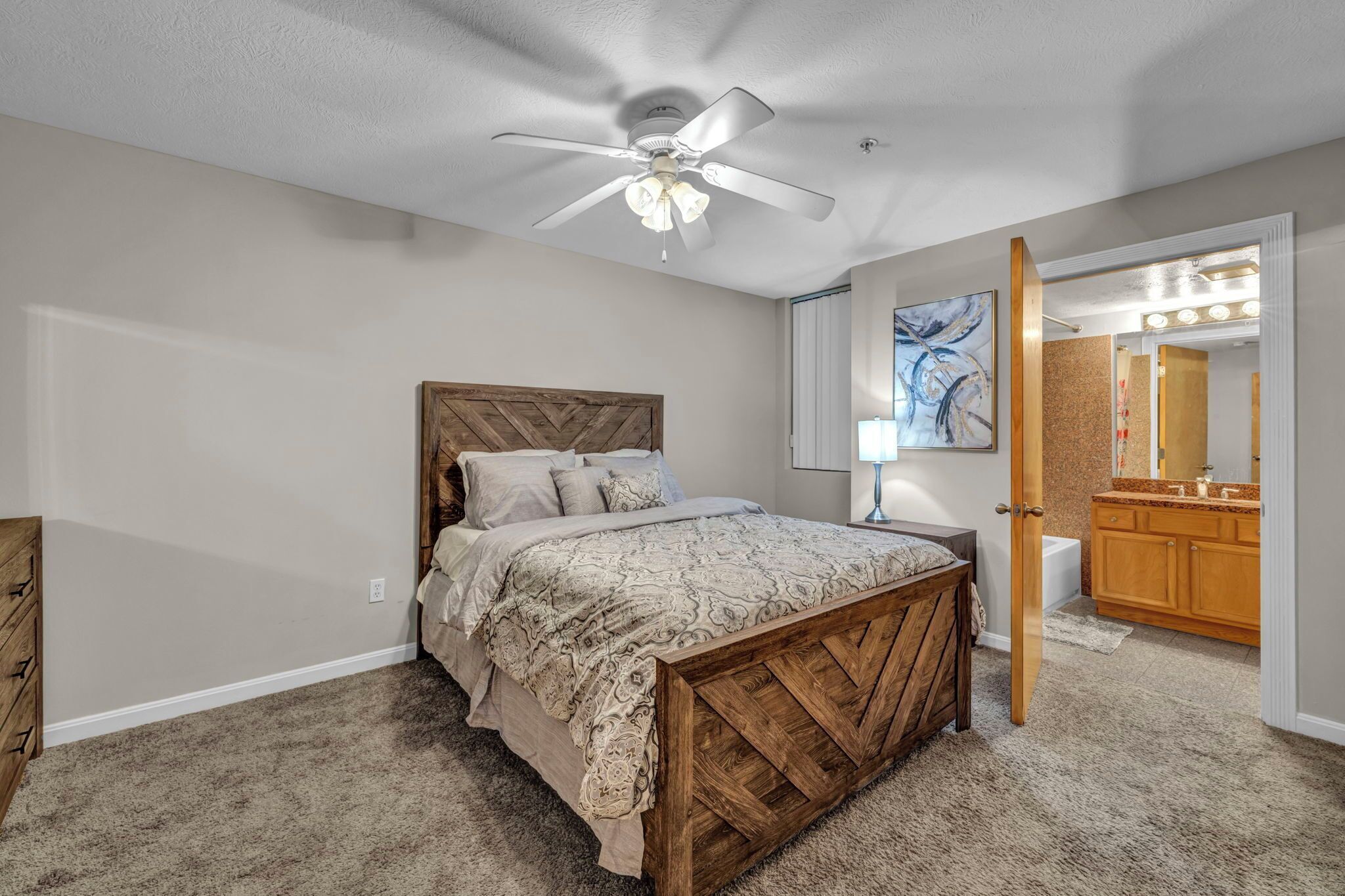 A bedroom with a wooden bed, a ceiling fan, a nightstand, a mirror, and a bathroom with a vanity and sink.