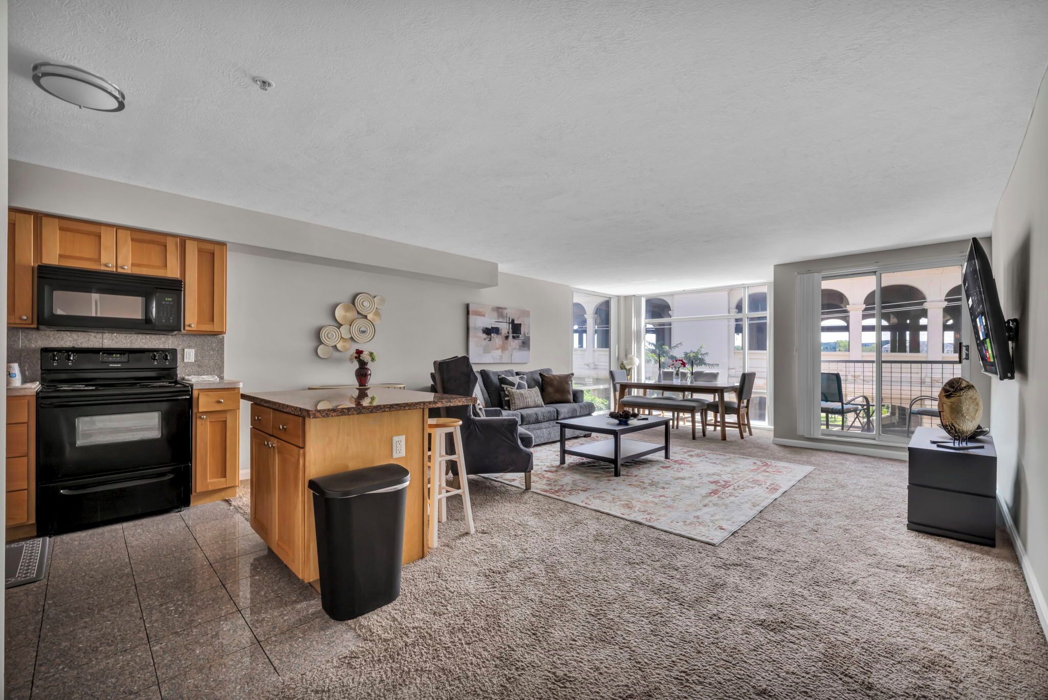 A modern kitchen with a built-in microwave and oven, a dining area with a table and chairs, a living room with a sofa and coffee table, and a large sliding glass door leading to an outdoor patio.