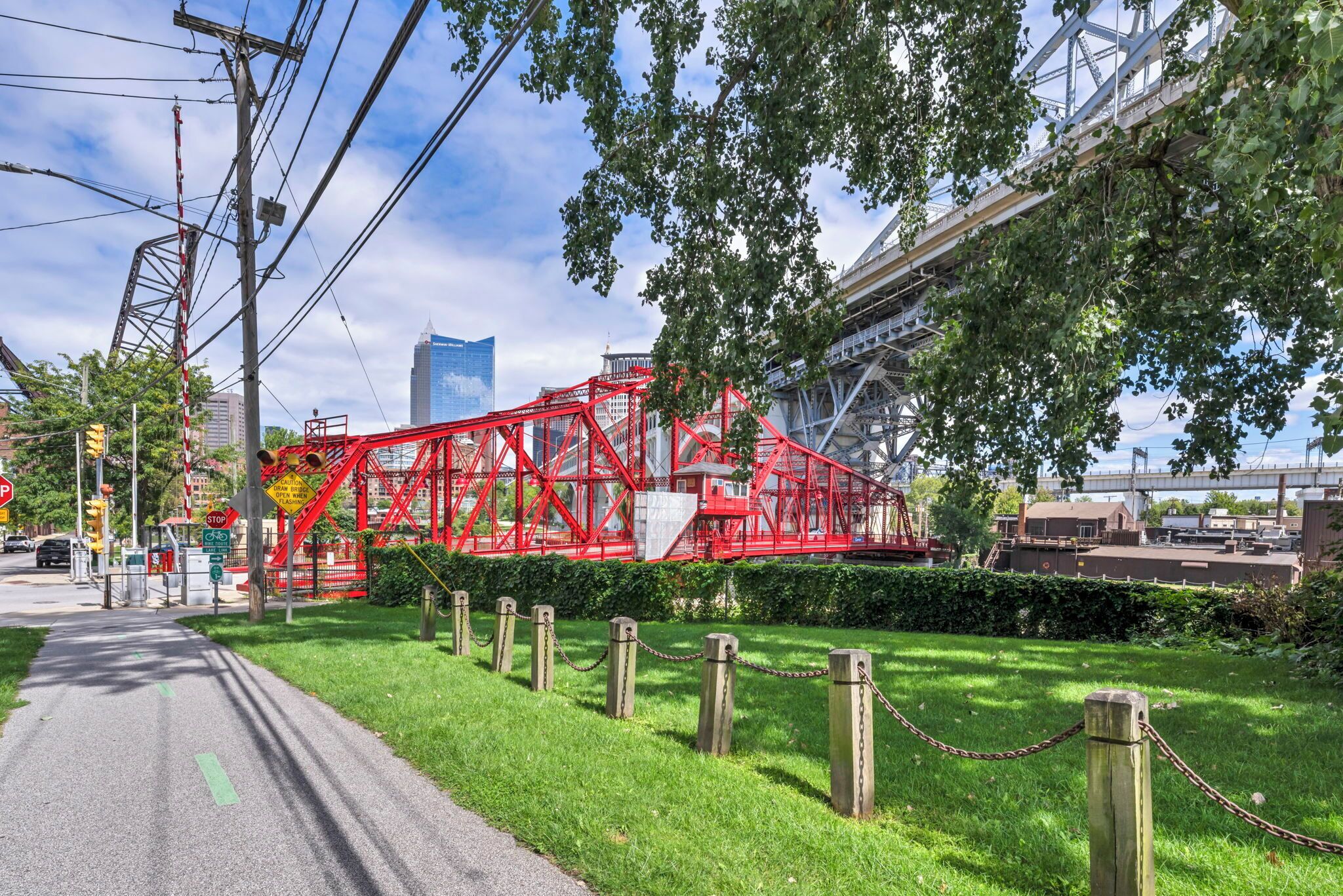 Een rode metalen constructie in een park met een verhard pad, een hek van gaas en een gebouw op de achtergrond.