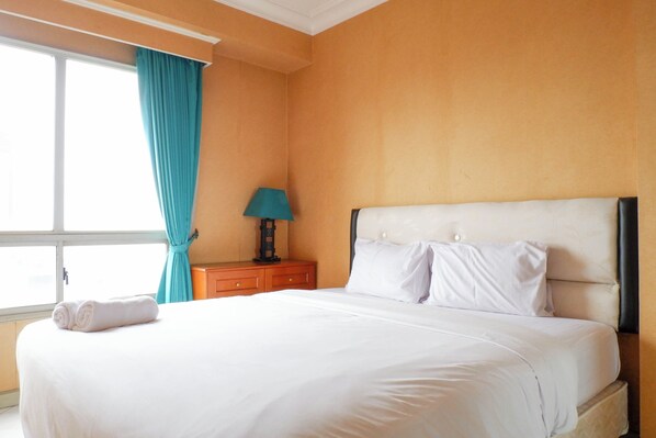 A neatly made bed with white linens, a teal curtain, a small bedside table with a lamp, and a window with curtains. - Best Deal 2Br Apartment At Taman Beverly (Surabaya)