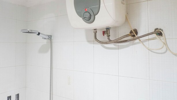 Ein Badezimmer mit weiß gefliesten Wänden, einer Duschbrause, einem Warmwasserbereiter und sichtbarer Sanitärinstallation.