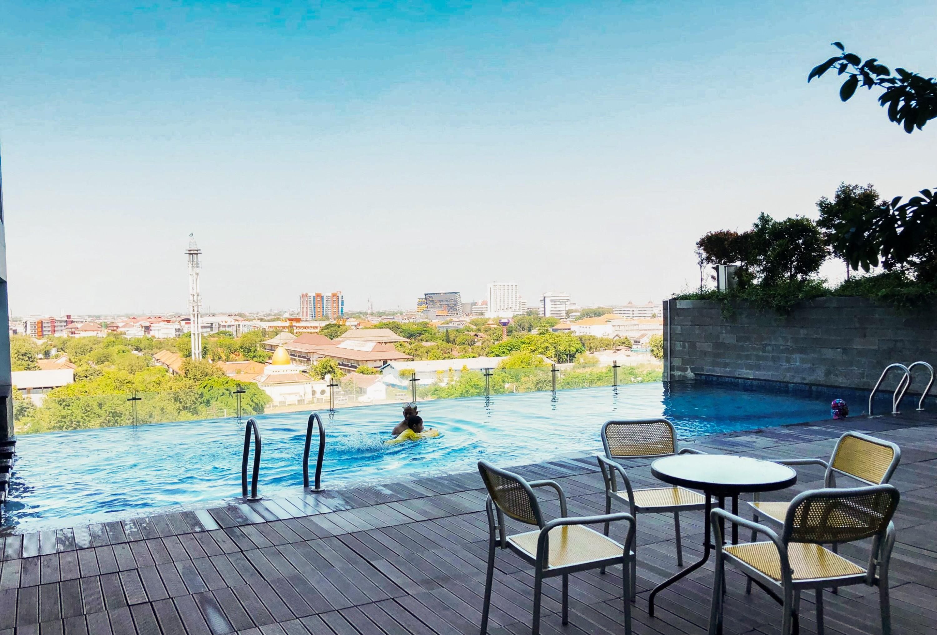 Una piscina infinita con vistas a un paisaje urbano, rodeada de una terraza de madera y mobiliario exterior.