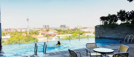 Une piscine à débordement offrant une vue sur le paysage urbain, entourée d’une terrasse en bois et d’un espace salon extérieur.