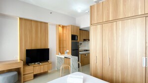 A hotel room with a bed, a desk with a TV, a kitchenette, and wooden cabinets. - Wonderful And Homey Studio Vasanta Innopark Apartment (Cikarang)