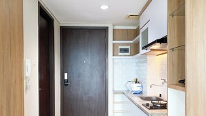 A modern kitchen with a dark wooden door, white cabinets, and a countertop with a sink and stove. - Wooden Style Studio At Lrt City Bekasi Apartment (Bekasi)