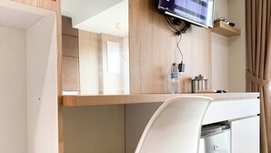 A modern hotel room with a wooden desk, a white chair, a flat-screen TV mounted on the wall, a water bottle, and a minibar. - Wooden Style Studio At Lrt City Bekasi Apartment (Bekasi)