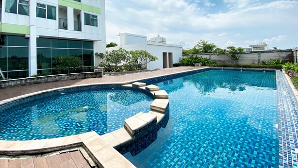 A modern building with a rooftop swimming pool, surrounded by greenery and a clear sky.
