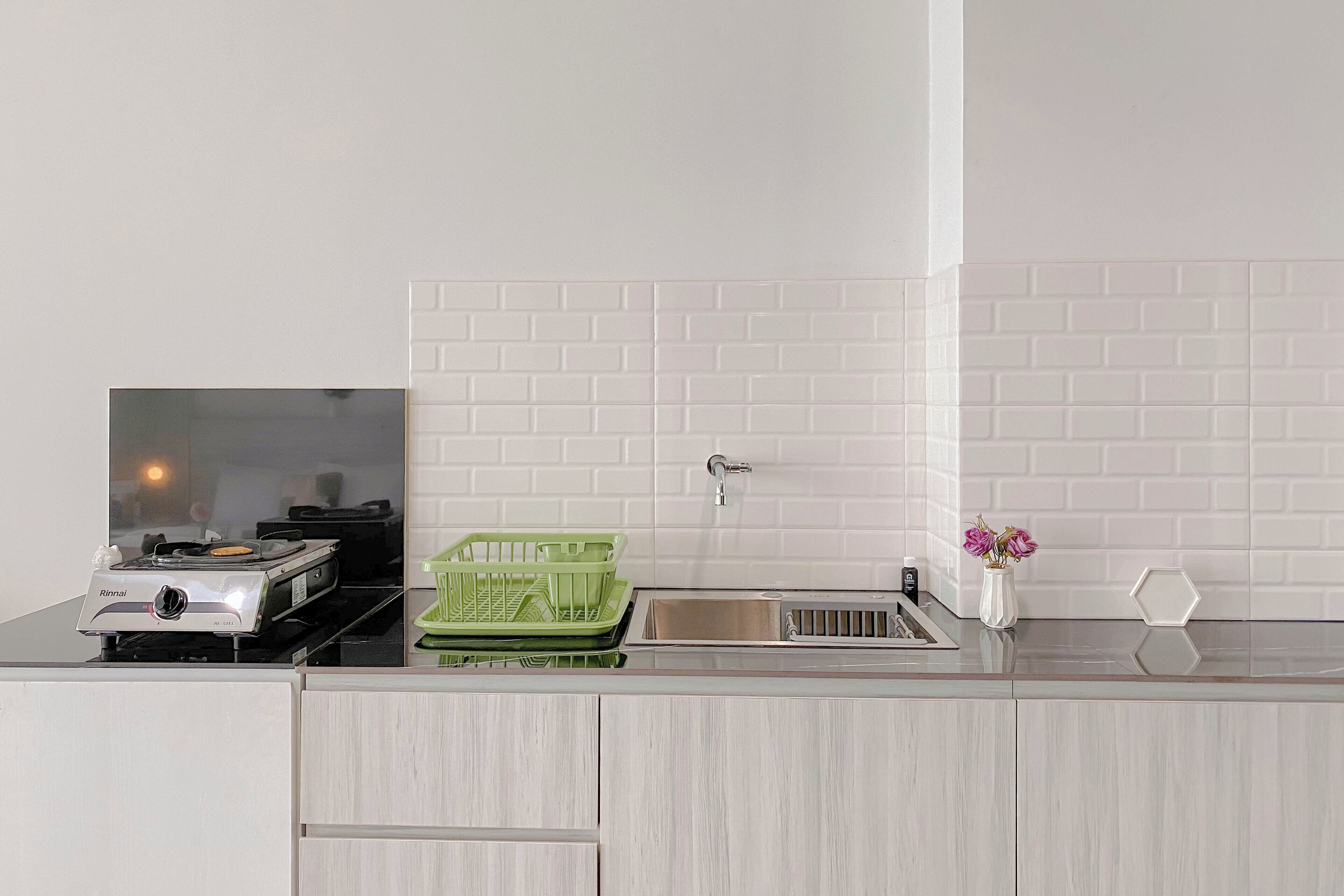 A kitchen with a white tiled backsplash, a sink, a green dish rack, a white vase with flowers, and a Rinnai brand stove.