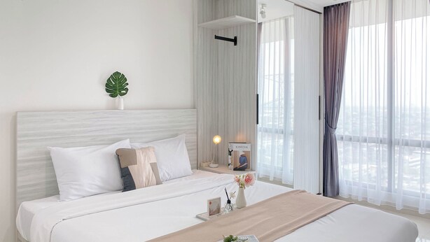 A modern hotel room with a large bed, a bedside table with a lamp, a plant, and a window with curtains.
