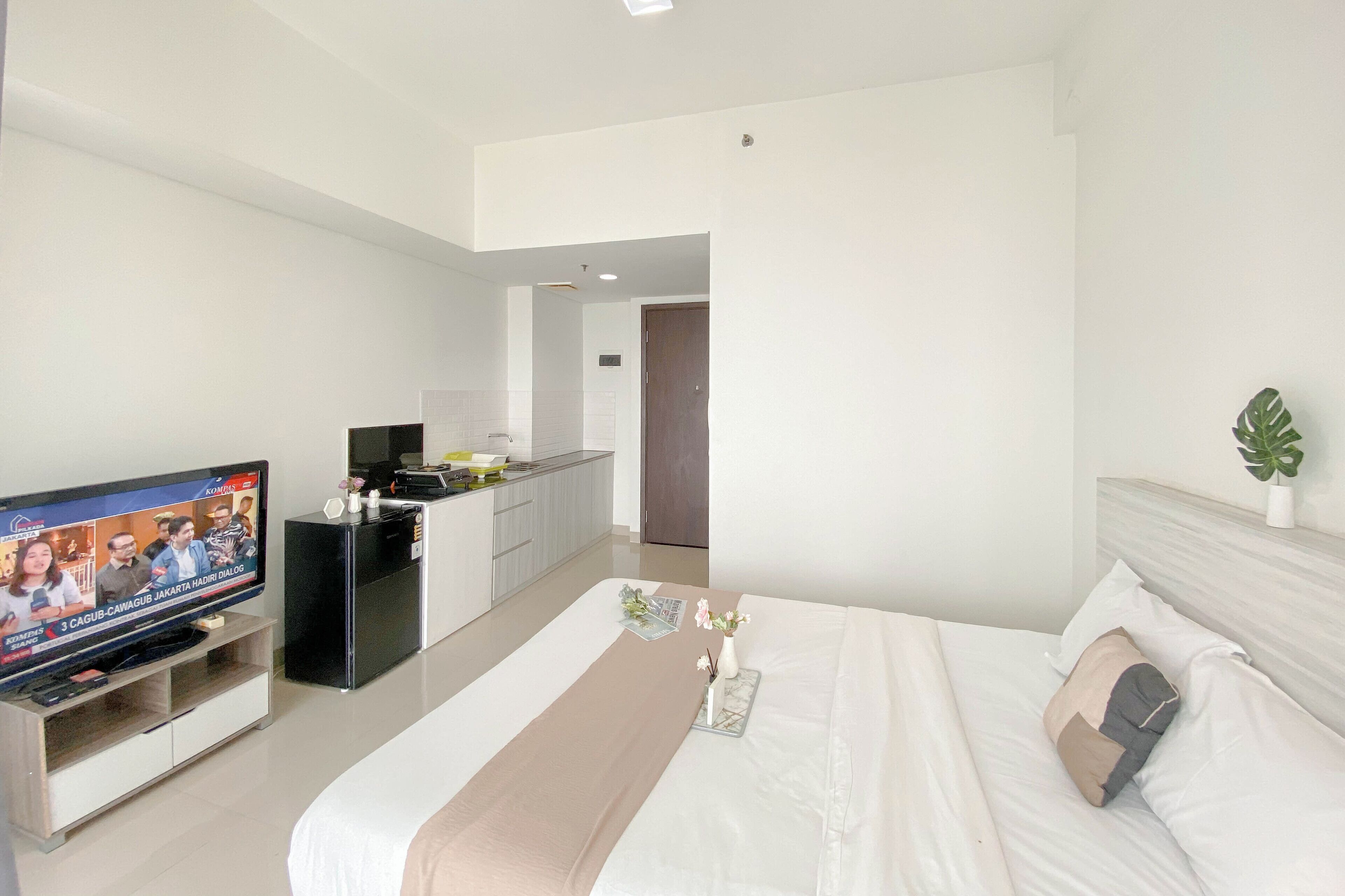 A modern hotel room with a large bed, a TV on a stand, a small kitchenette, and a door leading to another room.