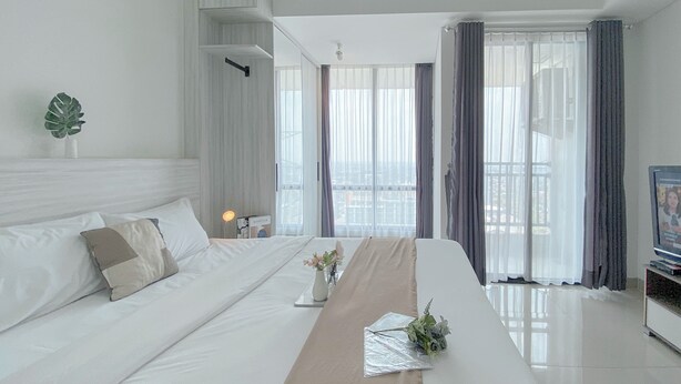 A modern hotel room with a large bed, a bedside table with a lamp, a view of the city through large windows, and a plant on the nightstand.