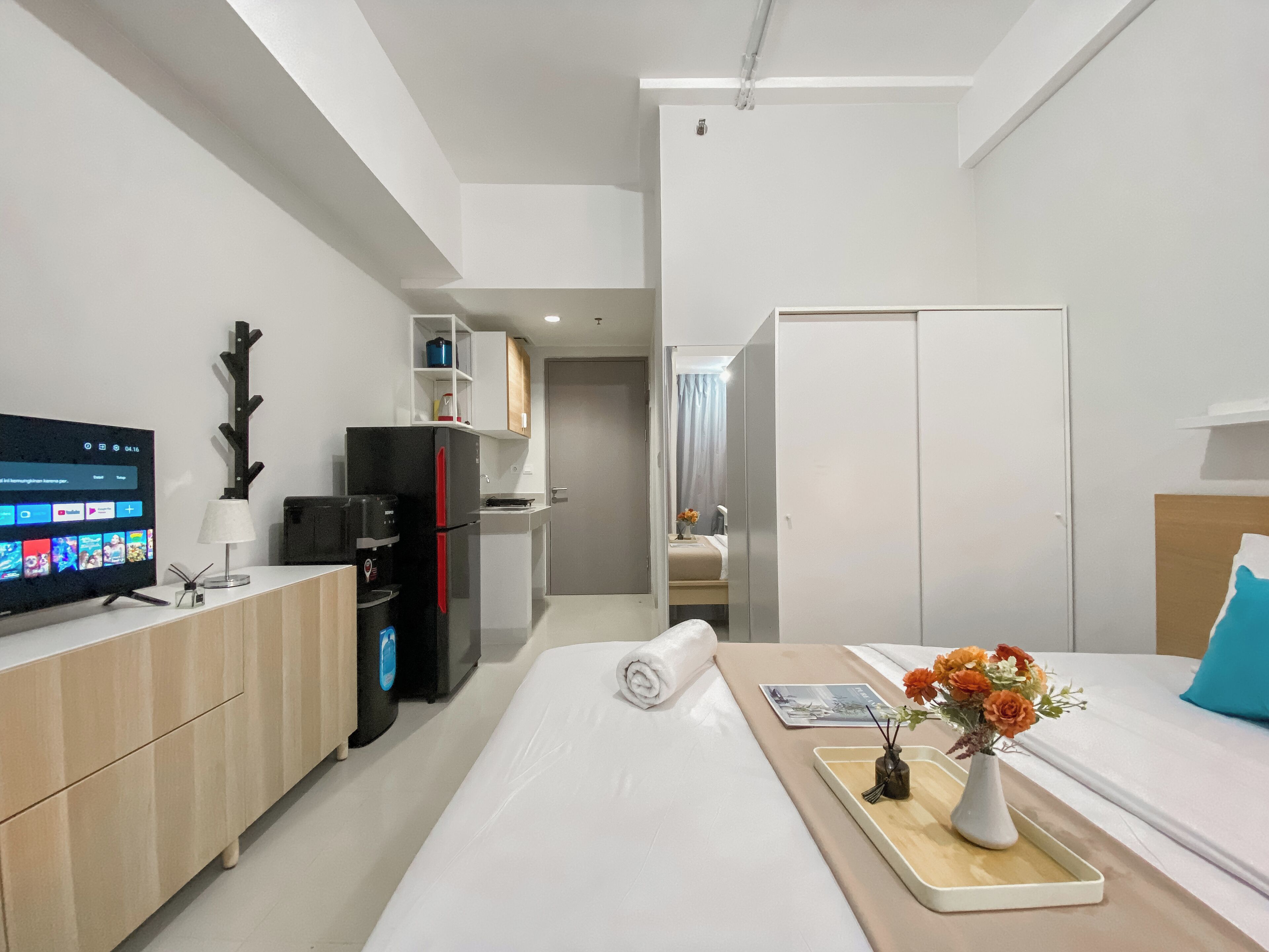 A modern hotel room with a kitchenette, a flat-screen TV, a bed with a tray and flowers, and a wardrobe.