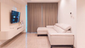 A modern living room with a flat-screen TV, a beige sectional sofa, and a large window with vertical blinds.