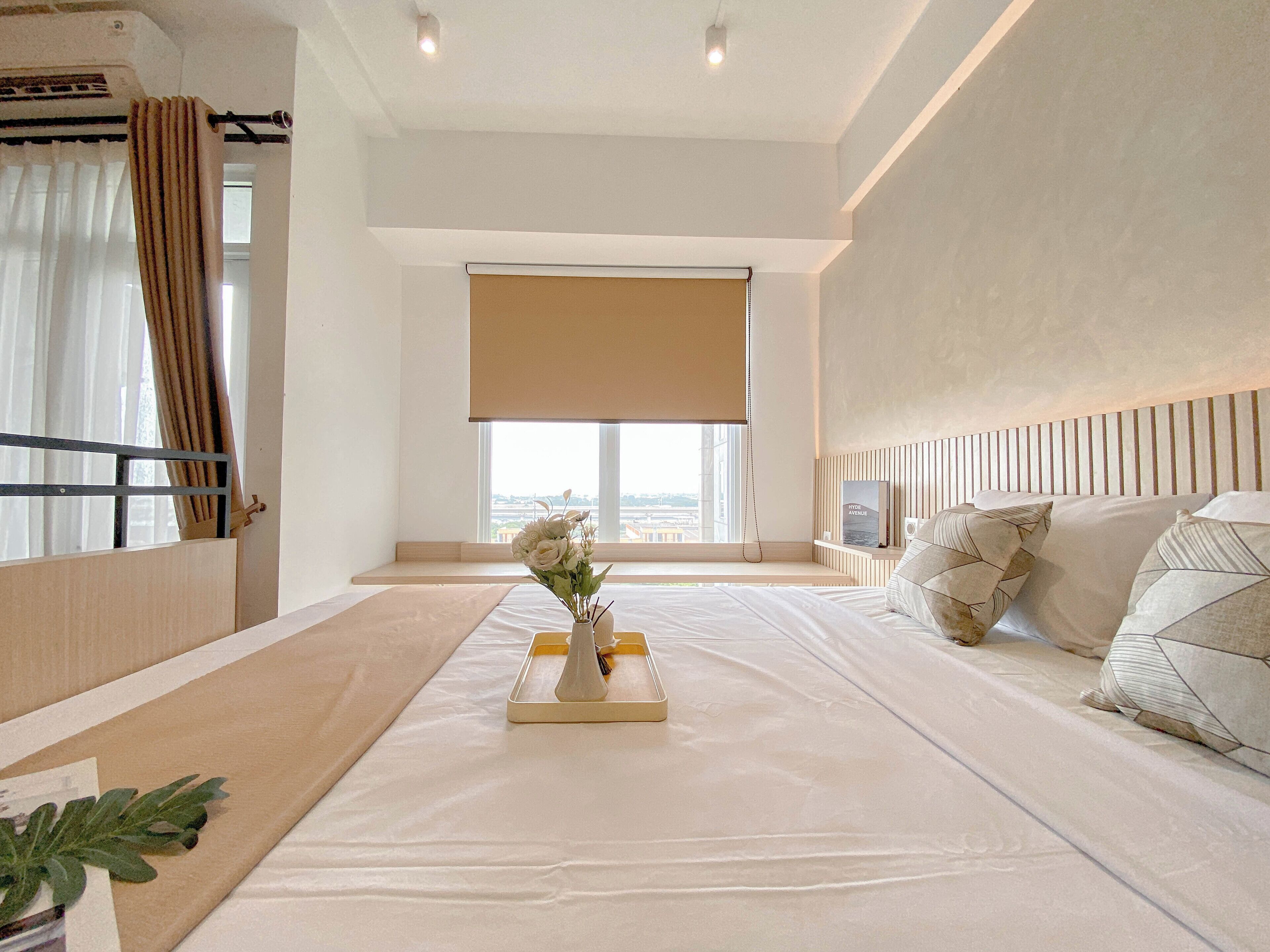 A modern hotel room with a large bed, a bedside table with a vase of flowers, a window with a view of the city, and a headboard with a built-in shelf.
