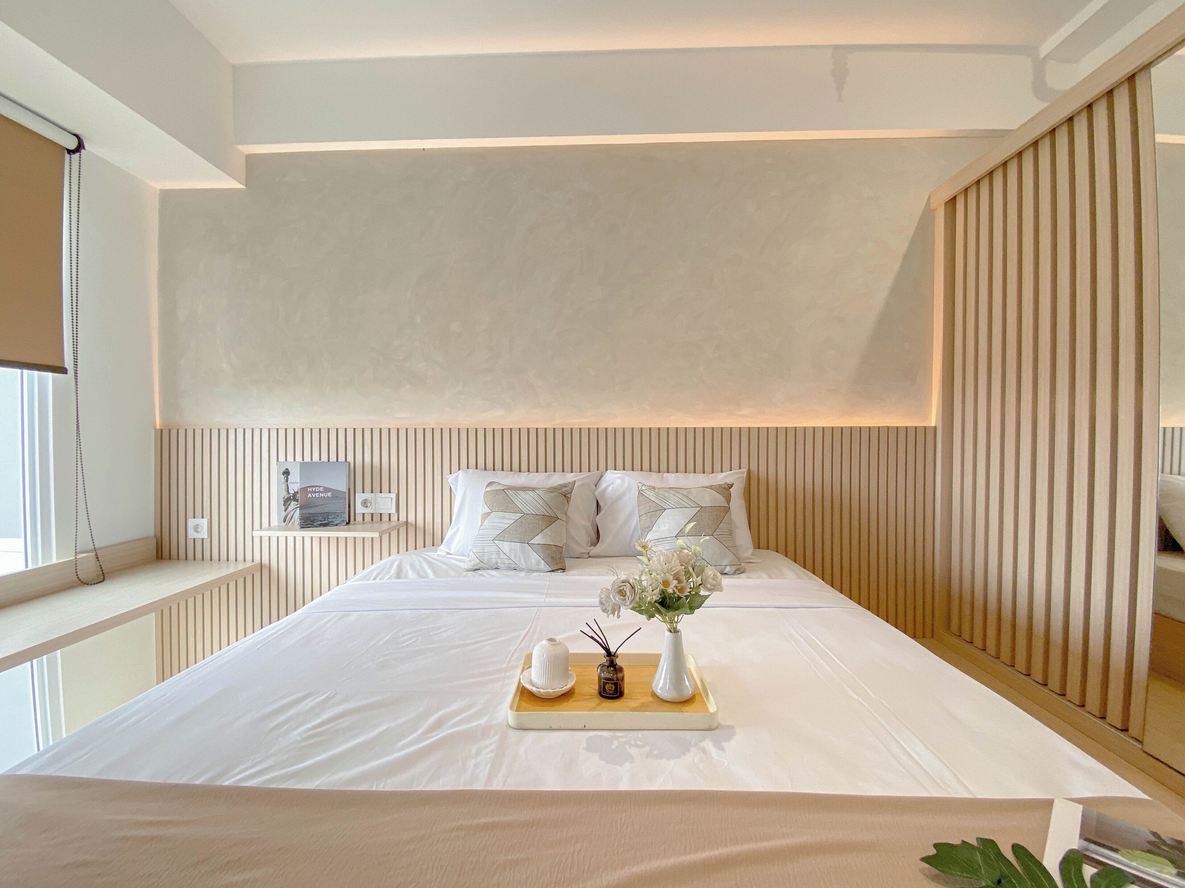A modern bedroom with a large bed, wooden paneling on the walls, a bedside table with a lamp, and a window with a curtain.