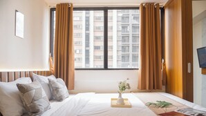 A hotel room with a large bed, a window with curtains, a vase with flowers, and a plant on the bedside table. - Spacious 3Br Apartment At Meikarta (Cikarang)
