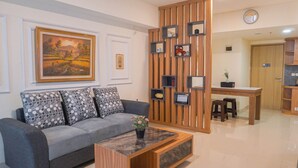 A modern living room with a grey sofa, a wooden coffee table, and a dining area with a wooden partition. - Spacious 3Br Apartment At Meikarta (Cikarang)
