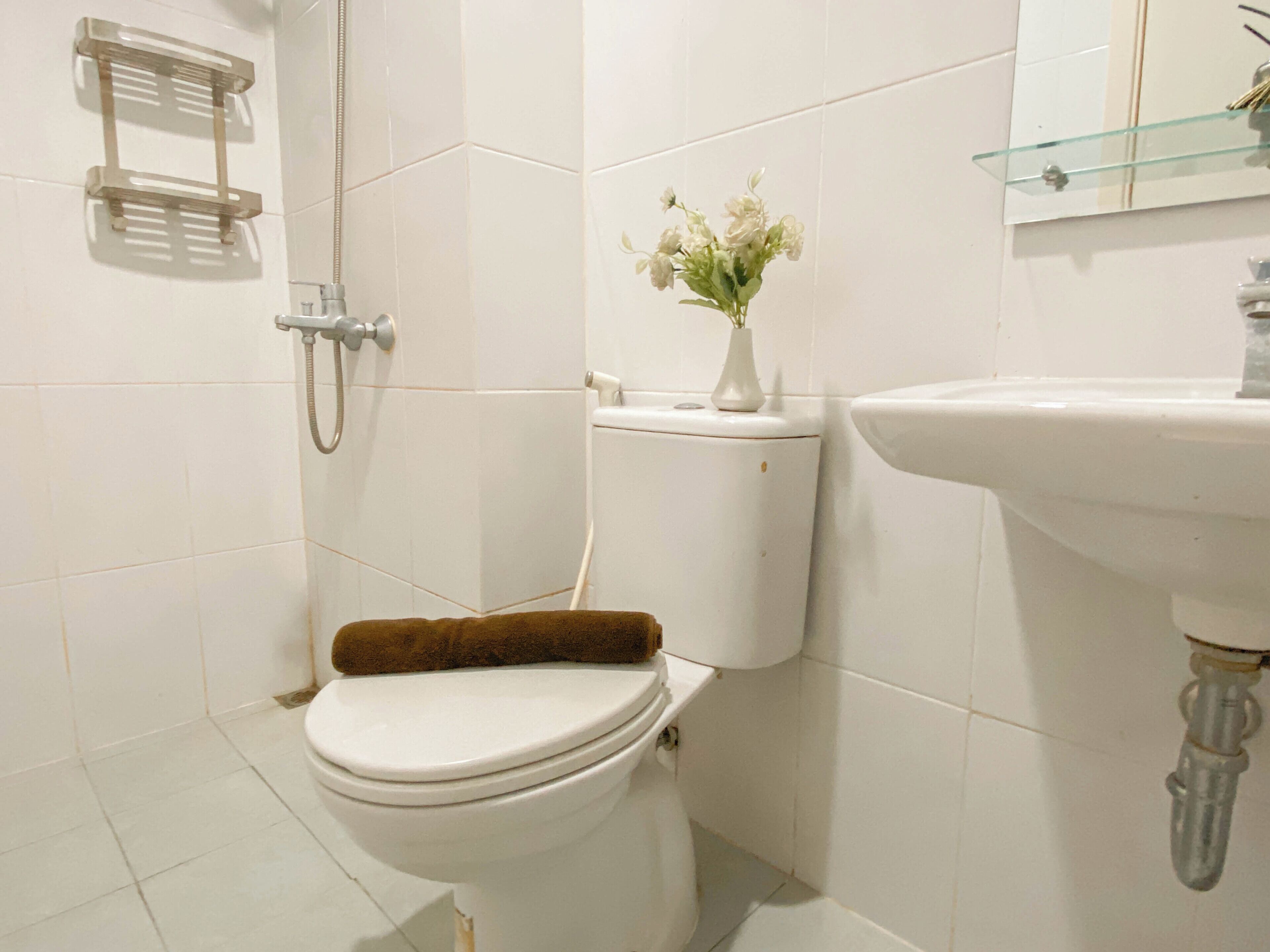 A bathroom with a toilet, a shower, a glass shelf with a vase of flowers, and a sink.