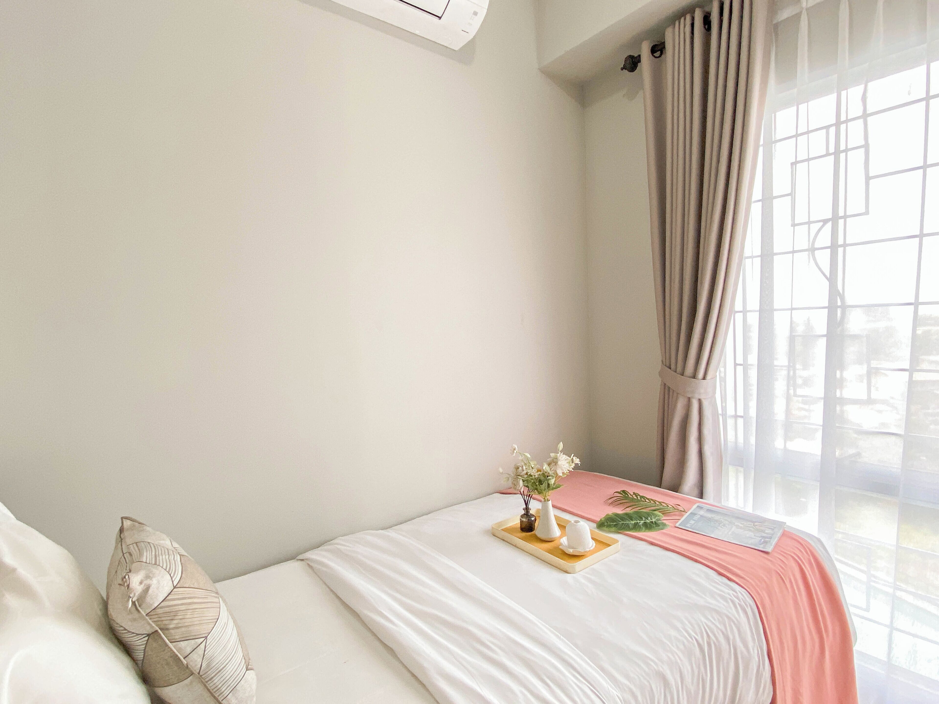 A neatly made bed with a pink blanket, a bedside table with a vase of flowers, and a window with curtains.