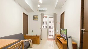 A modern living room with a sofa, a wooden coffee table, a TV, and a balcony with a view of buildings. - Big And Cozy 3Br Apartment At Transpark Juanda Bekasi Timur (Bekasi)