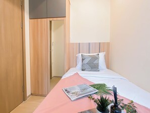 Une chambre à coucher avec un lit, un magazine posé sur le lit, une couvre-lit rose, une plante et une porte donnant sur une autre pièce.