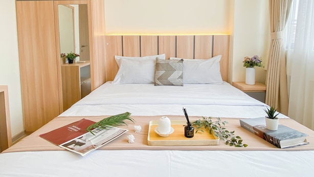 A neatly made bed with white linens, a bedside table with a book and a small potted plant, and a wooden wardrobe with a mirror. - Warm 2Br Apartment At Meikarta (Cikarang)