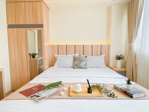 A neatly made bed with white linens, a bedside table with a book and a small potted plant, and a wooden wardrobe with a mirror. - Warm 2Br Apartment At Meikarta (Cikarang)