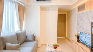 A modern living room with a sofa, a coffee table with a plant, a TV on a wooden stand, and an air conditioning unit on the ceiling. - Warm 2Br Apartment At Meikarta (Cikarang)