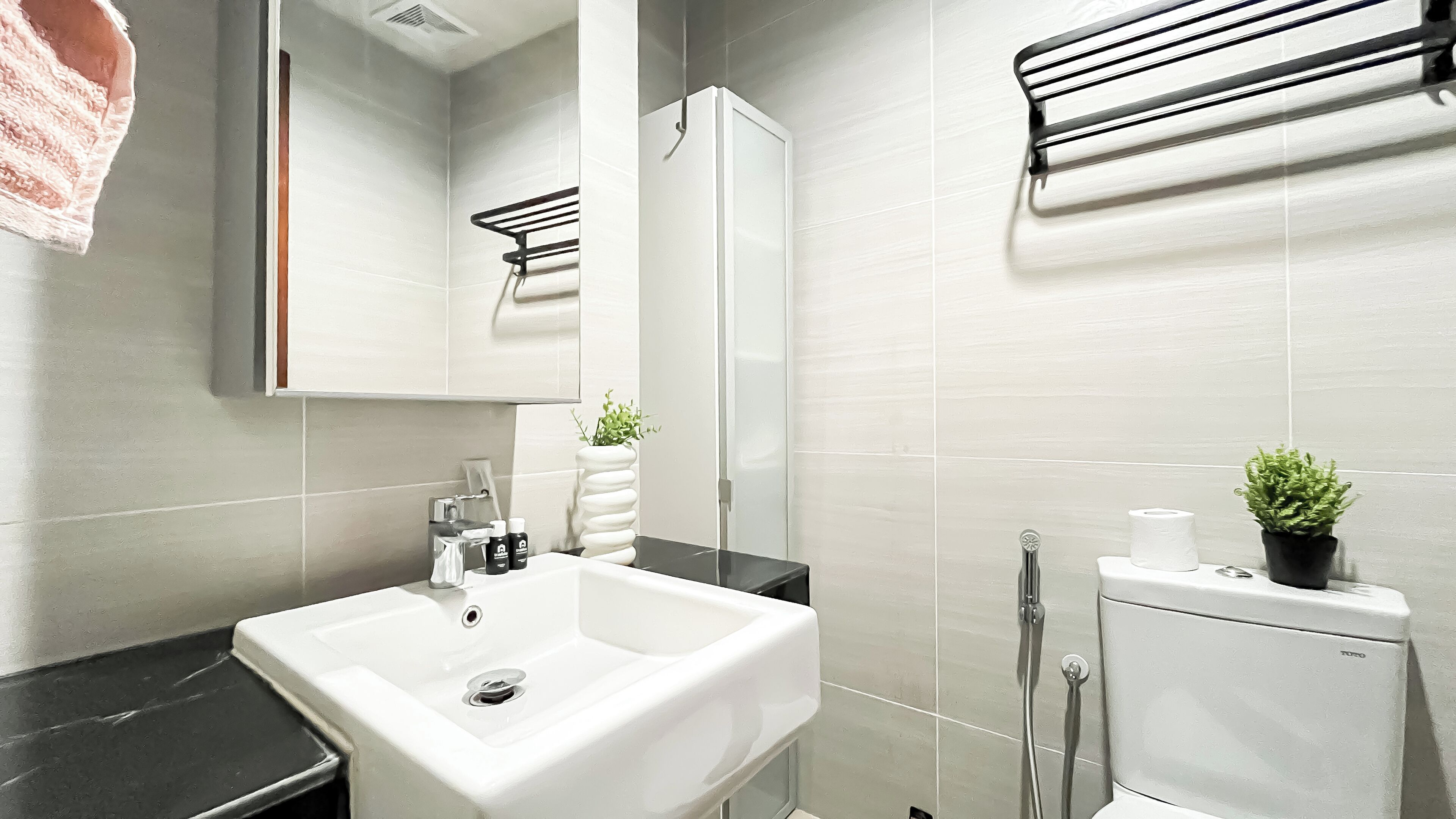 A modern bathroom with a white sink, black countertop, and a toilet. There is a towel rack above the sink and a small potted plant on the toilet.