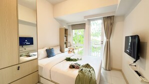 A hotel room with a bed, a TV, a desk with a computer, a wardrobe, and a window with a view of greenery. - Basic And Compact Studio At Tokyo Riverside Pik 2 Apartment (Teluknaga)