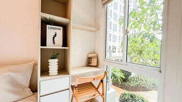 Una habitación con un escritorio blanco, una silla, una estantería con un libro titulado 'LONDON' y una ventana que ofrece vistas a zonas verdes y edificios.